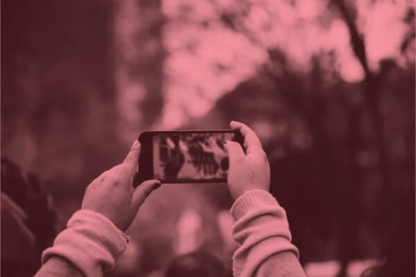 Someone holds up a smartphone to record or photograph a scene where a crowd of people has gathered. The image has a reddish-pink tonal filter applied to it. The person filming is in the foreground, their hands and device in focus, while the crowd behind them appears blurred. The scene suggests bystander documentation of a public event or gathering, capturing the modern phenomenon of people recording moments through their devices rather than simply observing directly.