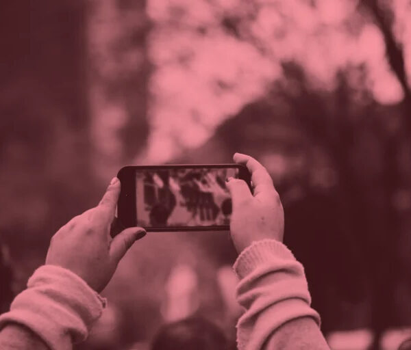 Someone holds up a smartphone to record or photograph a scene where a crowd of people has gathered. The image has a reddish-pink tonal filter applied to it. The person filming is in the foreground, their hands and device in focus, while the crowd behind them appears blurred. The scene suggests bystander documentation of a public event or gathering, capturing the modern phenomenon of people recording moments through their devices rather than simply observing directly.