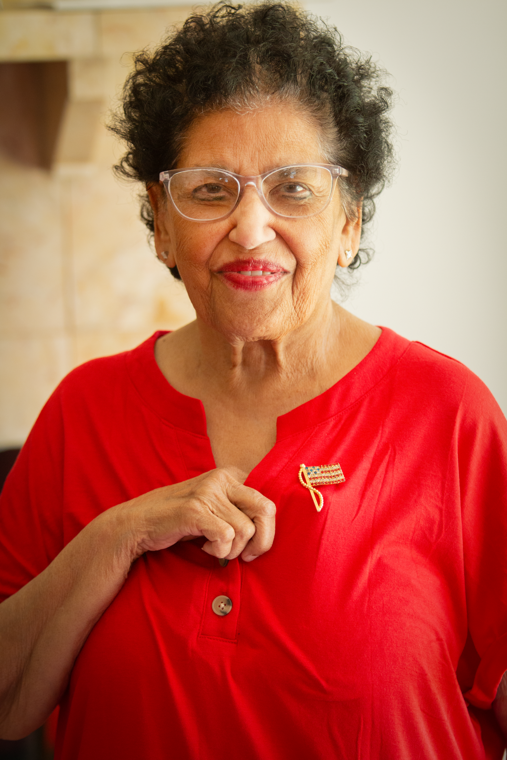 Amna Zahir, an Indian woman with short black hair wearing a red shirt shows off her American flag lapel pin that is fastened to her shirt.