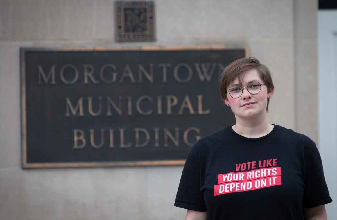 Young person with short brown hair and round glasses wearing black t-shirt with red text "VOTE LIKE YOUR RIGHTS DEPEND ON IT" standing in front of "MORGANTOWN MUNICIPAL BUILDING" sign on white wall.