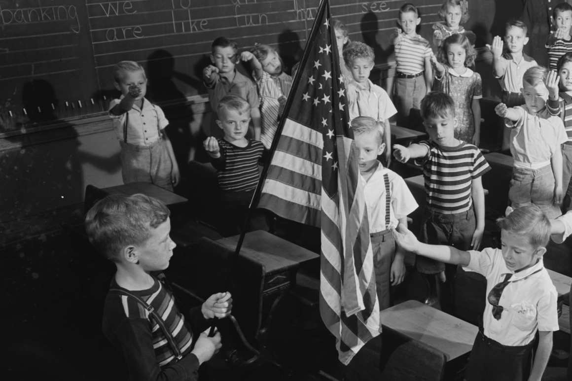 Black and white historical photograph of children in classroom setting, with one child standing near American flag while others gather around, framed pictures visible on walls and large windows in background.