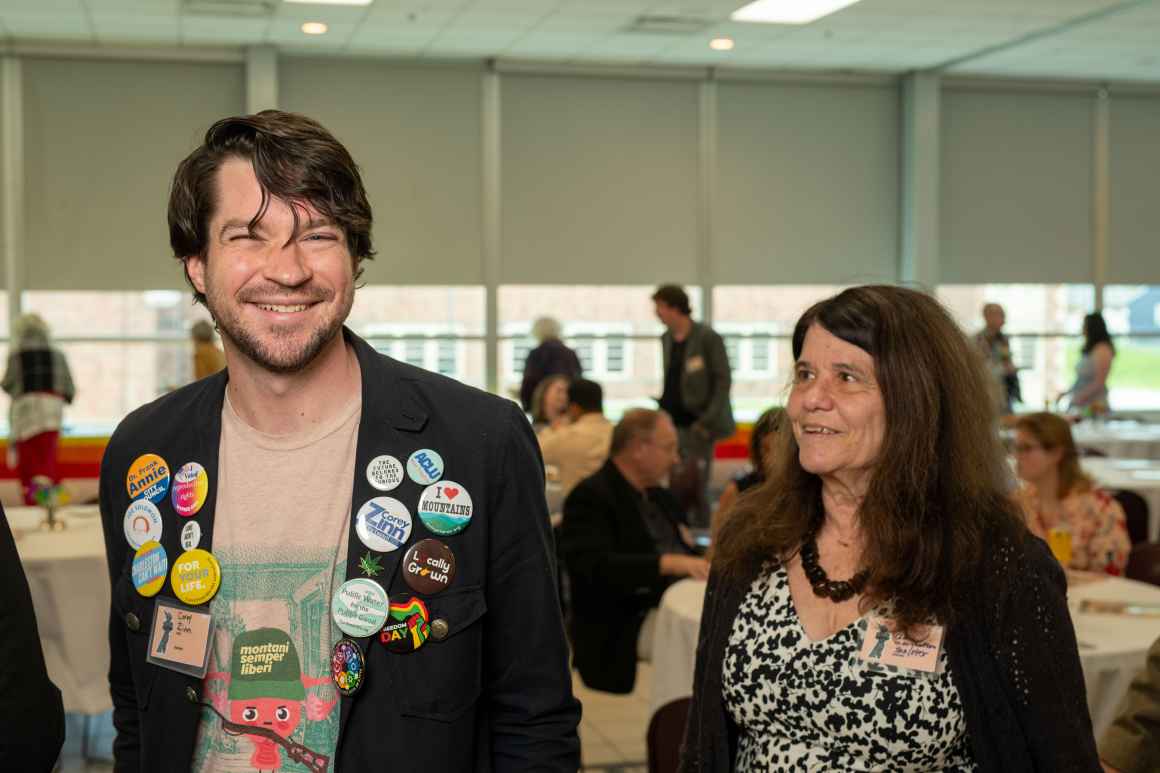 A man with several buttons on his jacket poses for a picture while a friend of his looks on.