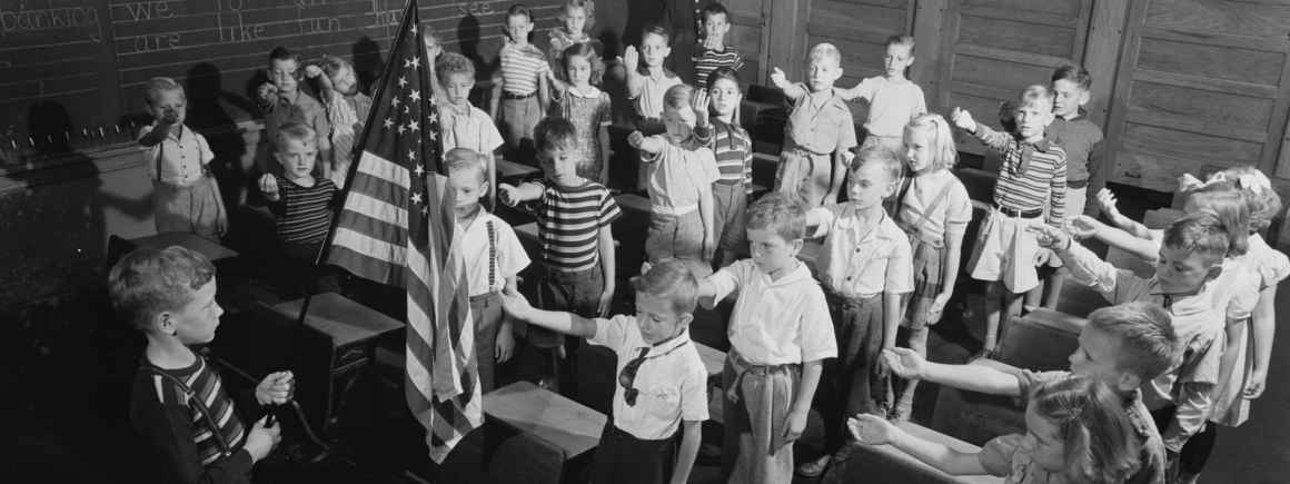 Black and white vintage photograph of elementary school classroom with approximately 30 children standing and raising their right hands, presumably reciting Pledge of Allegiance, with American flag visible and chalkboard in background.