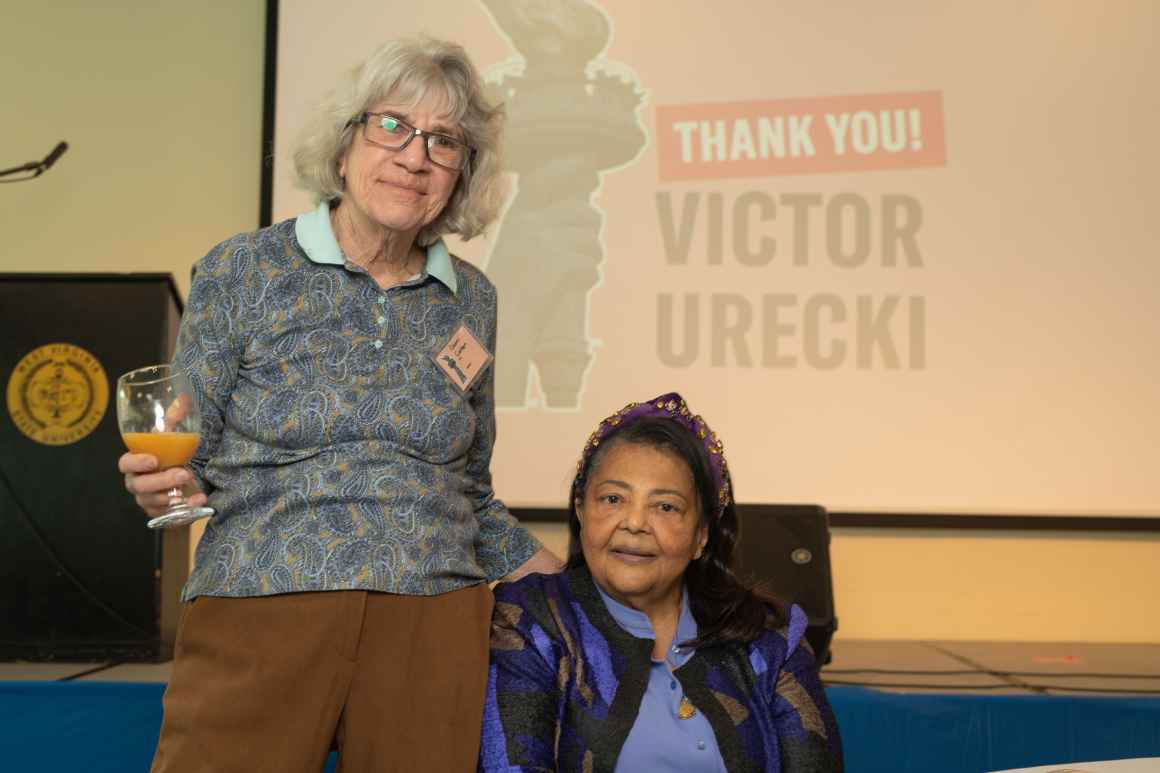 A woman who is standing is holding a glass of orange juice next to a woman in a purple and black jacket who is seated.