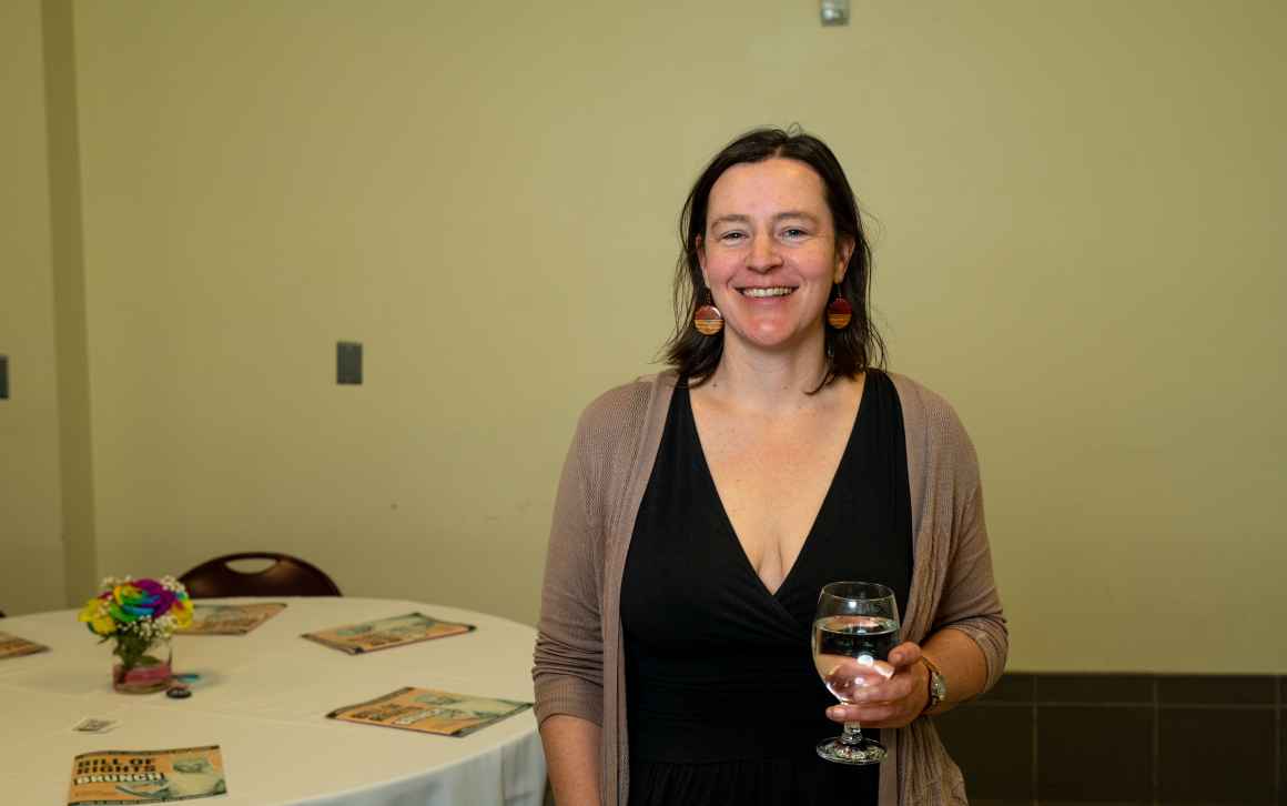 A woman in a black dress and tan jacket holds a glass of water and poses for a picture.