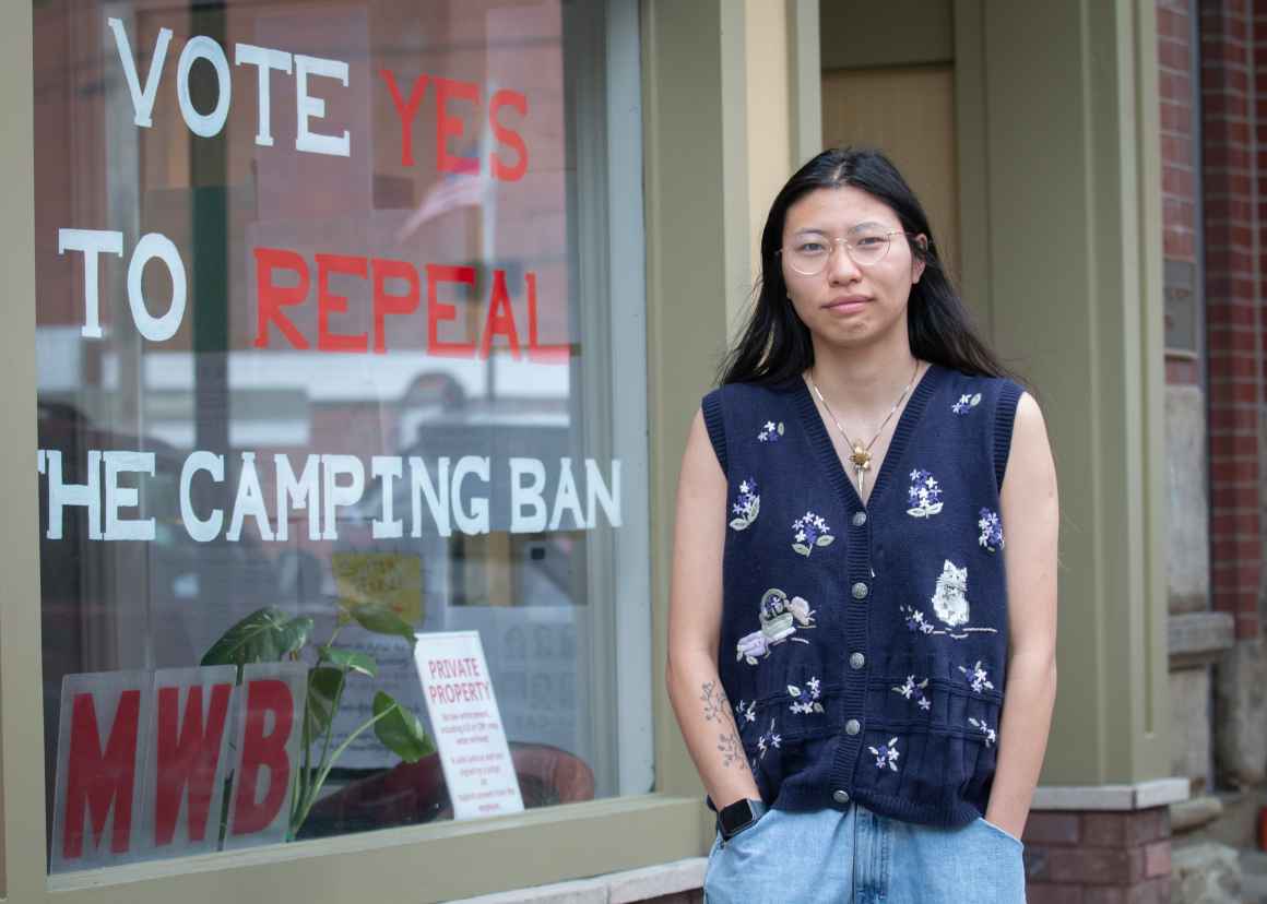 A young person in a navy floral cardigan and glasses standing in front of storefront window displaying "VOTE YES TO REPEAL THE CAMPING BAN" sign, with brick buildings visible in background.