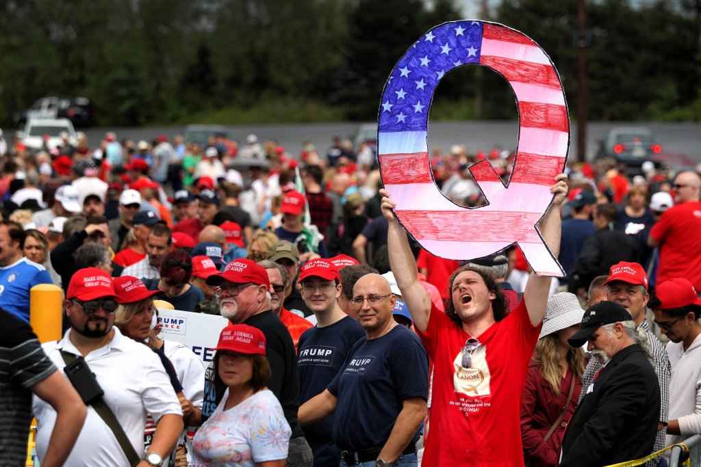A white man with long hair hold up a large "Q" painted like the American flag for the conspiracy theory Q Anon. 