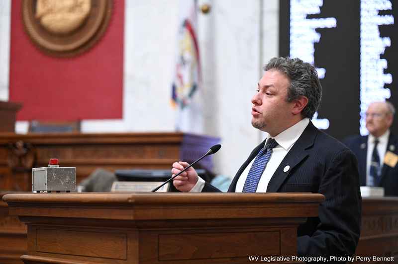 Eli Baumwell speaks at the West Virginia Legislature wearing a dark suit and blue tie. 