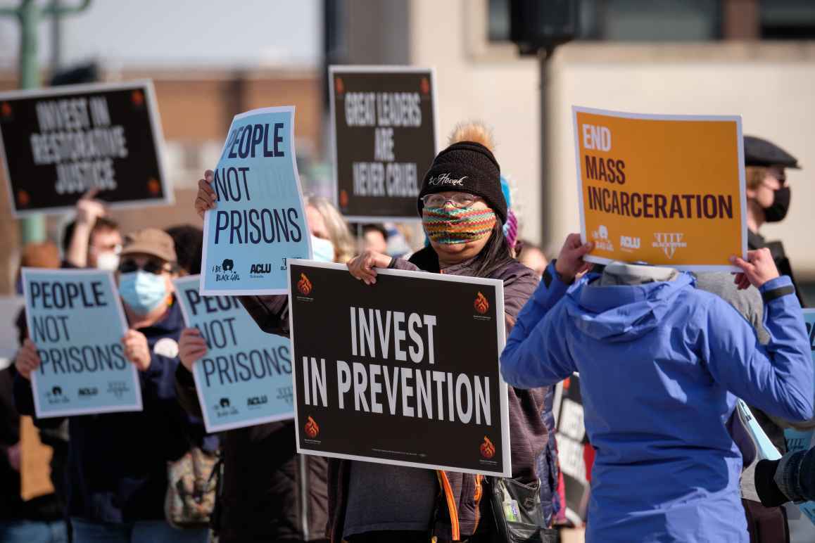 Protesters wave signs saying "INVEST IN PREVENTION," "PEOPLE NOT PRISONS," AND "END MASS INCARCERATION."