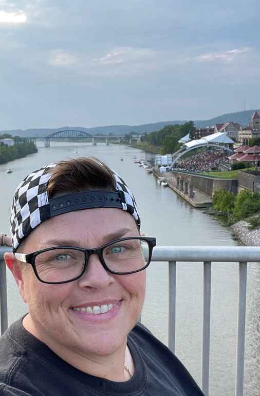 Miranda Johnson, a white woman with dark glasses, short hair and a backwards checker-print baseball hat, smiles for the camera with downtown Charleston's waterfront in the background. 