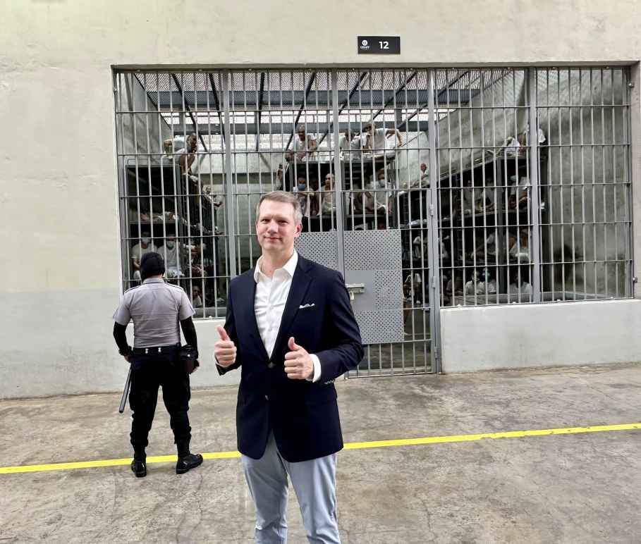 Riley Moore, a Congressman, stands in a suit giving a thumbs up in front of a large prison cell holding numerous people being watched over by a guard