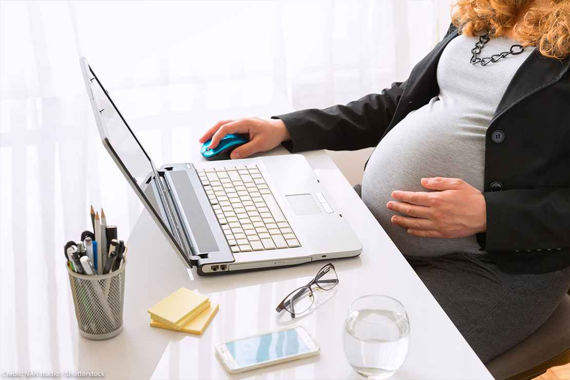 A pregnant worker touches their belly while performing work duties on a laptop