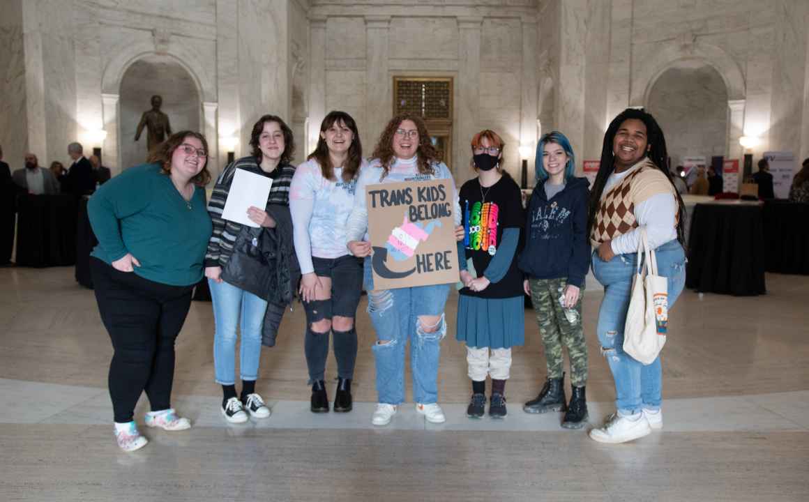 Seven attendees of the Public Hearing on HB 2007 pose with a sign that reads "Trans Kids Belong Here and a picture of West Virginia"