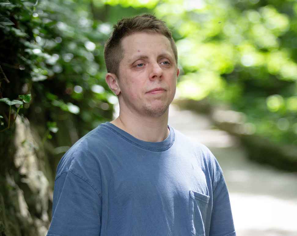 Robb Livingood, a white man with short light brown hair and a tattoo sleeve on his left arm, is pictured in a light blue shirt standing along a nature trail.