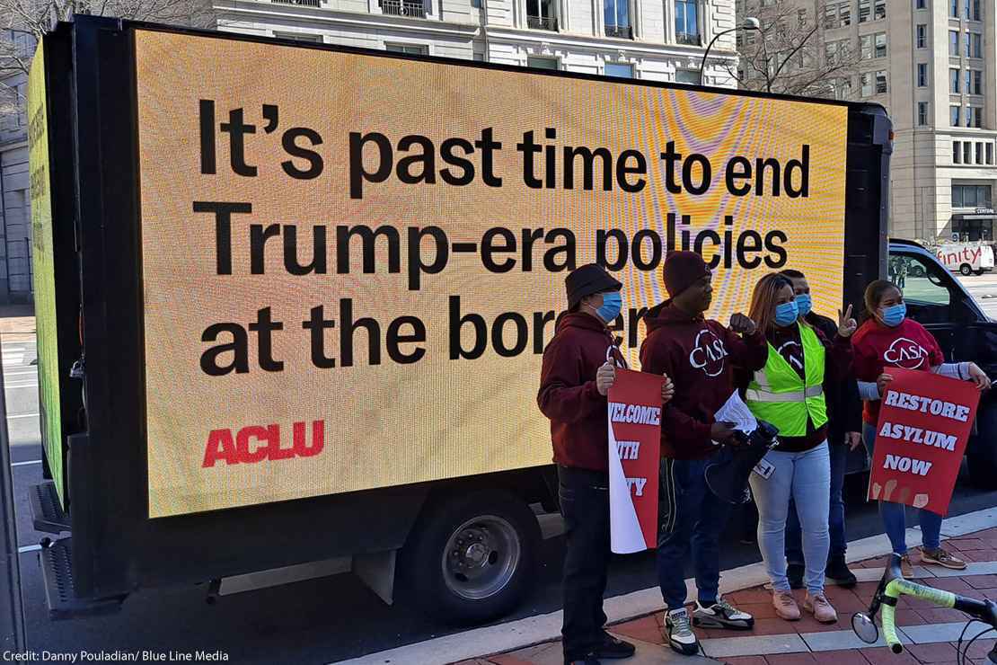 Protestors stand in front of a digital sign that says "It's past time to end Trump-era policies at the border"