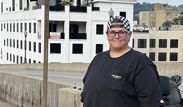 Miranda Johnson, a white woman with dark glasses, short hair and a backwards checker-print baseball hat, smiles for the camera with downtown Charleston's waterfront in the background. 