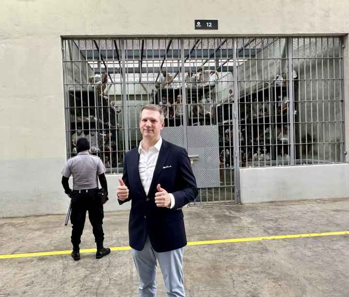 Riley Moore, a Congressman, stands in a suit giving a thumbs up in front of a large prison cell holding numerous people being watched over by a guard