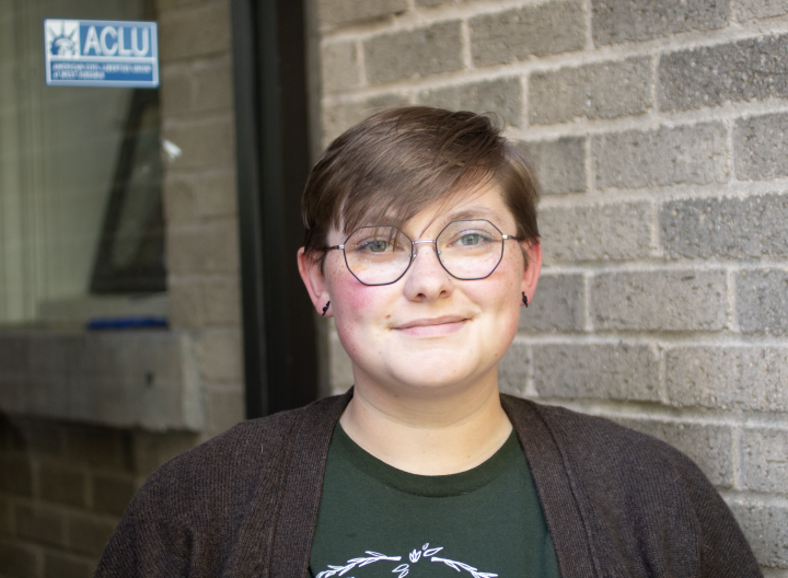 Sam Green is a white person with short medium brown hair, blue eyes and glasses, wearing a green shirt and brown sweater, standing outside of the ACLU offices.
