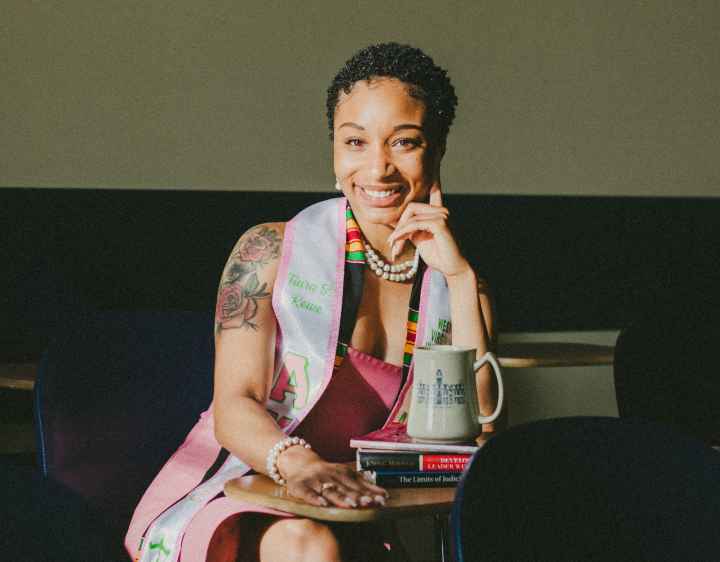 Tiara Rowe is a young Black women with short hair and a shoulder tattoo sits at a table with books and a coffee mug