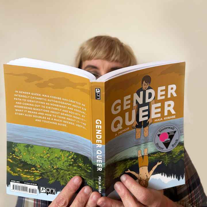 A white person with blond hair and glasses reads a copy of "Gender Queer" against a solid white backdrop