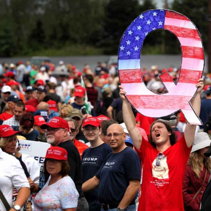 A white man with long hair hold up a large "Q" painted like the American flag for the conspiracy theory Q Anon. 