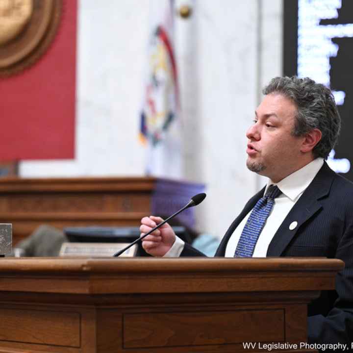 Eli Baumwell speaks at the West Virginia Legislature wearing a dark suit and blue tie. 