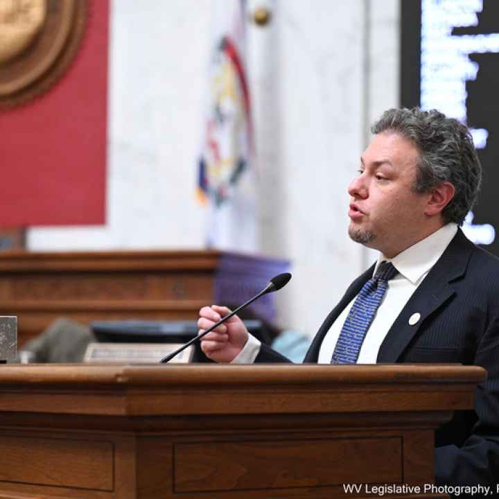 Eli Baumwell speaks at the West Virginia Legislature wearing a dark suit and blue tie.