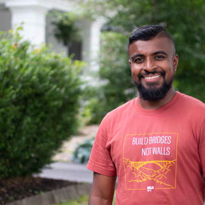 Kasun Wijayagurusinghe is pictured in an orange Build Bridges Not Walls tee shirt in front of the ACLU-WV Charleston Office