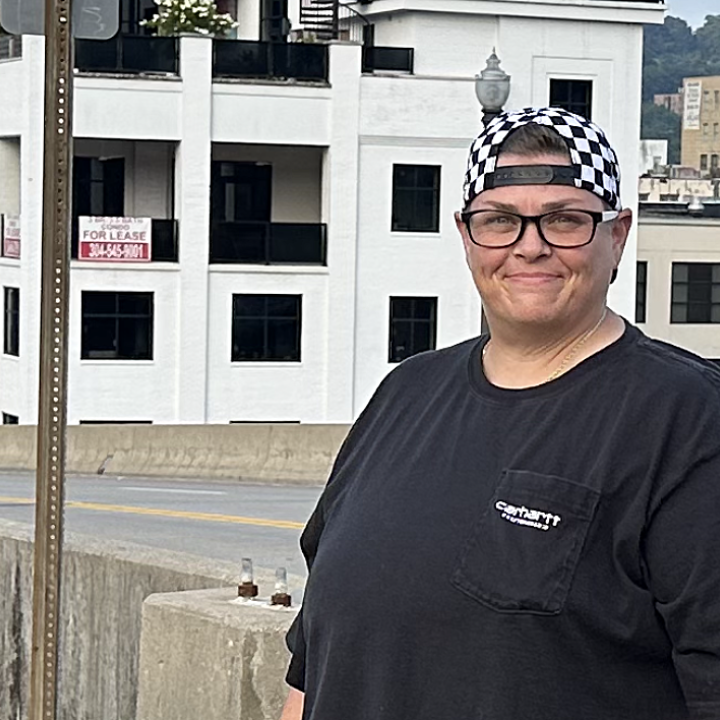 Miranda Johnson, a white woman with dark glasses, short hair and a backwards checker-print baseball hat, smiles for the camera with downtown Charleston's waterfront in the background. 