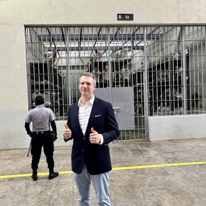 Riley Moore, a Congressman, stands in a suit giving a thumbs up in front of a large prison cell holding numerous people being watched over by a guard