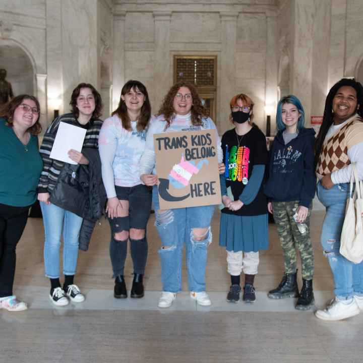 Seven attendees of the Public Hearing on HB 2007 pose with a sign that reads "Trans Kids Belong Here and a picture of West Virginia"