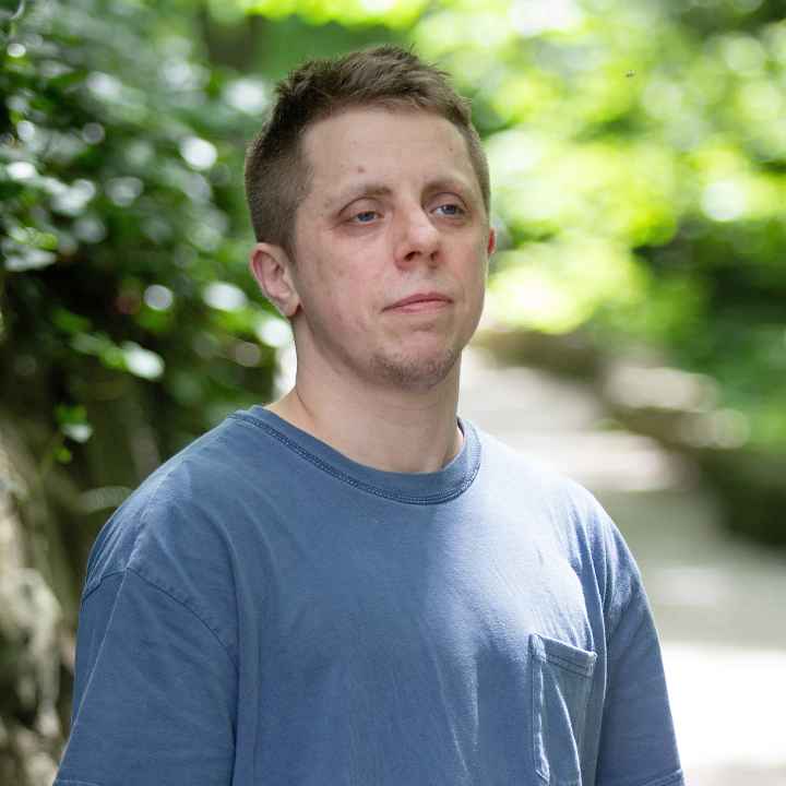 Robb Livingood, a white man with short light brown hair and a tattoo sleeve on his left arm, is pictured in a light blue shirt standing along a nature trail.