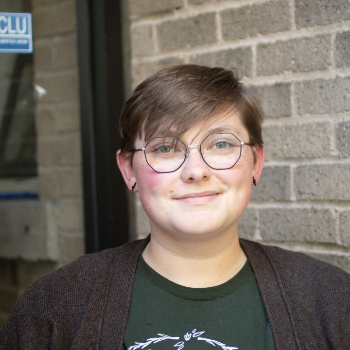 Sam Green is a white person with short medium brown hair, blue eyes and glasses, wearing a green shirt and brown sweater, standing outside of the ACLU offices.