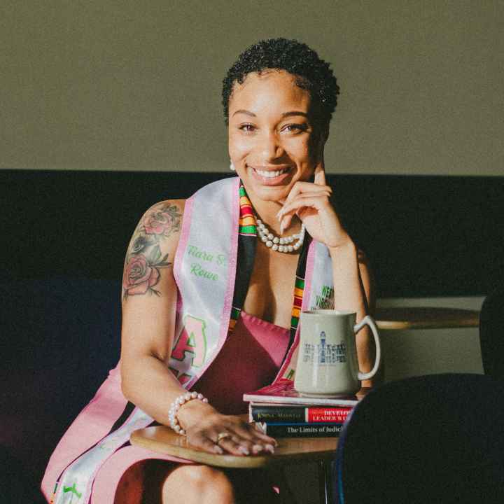 Tiara Rowe is a young Black women with short hair and a shoulder tattoo sits at a table with books and a coffee mug